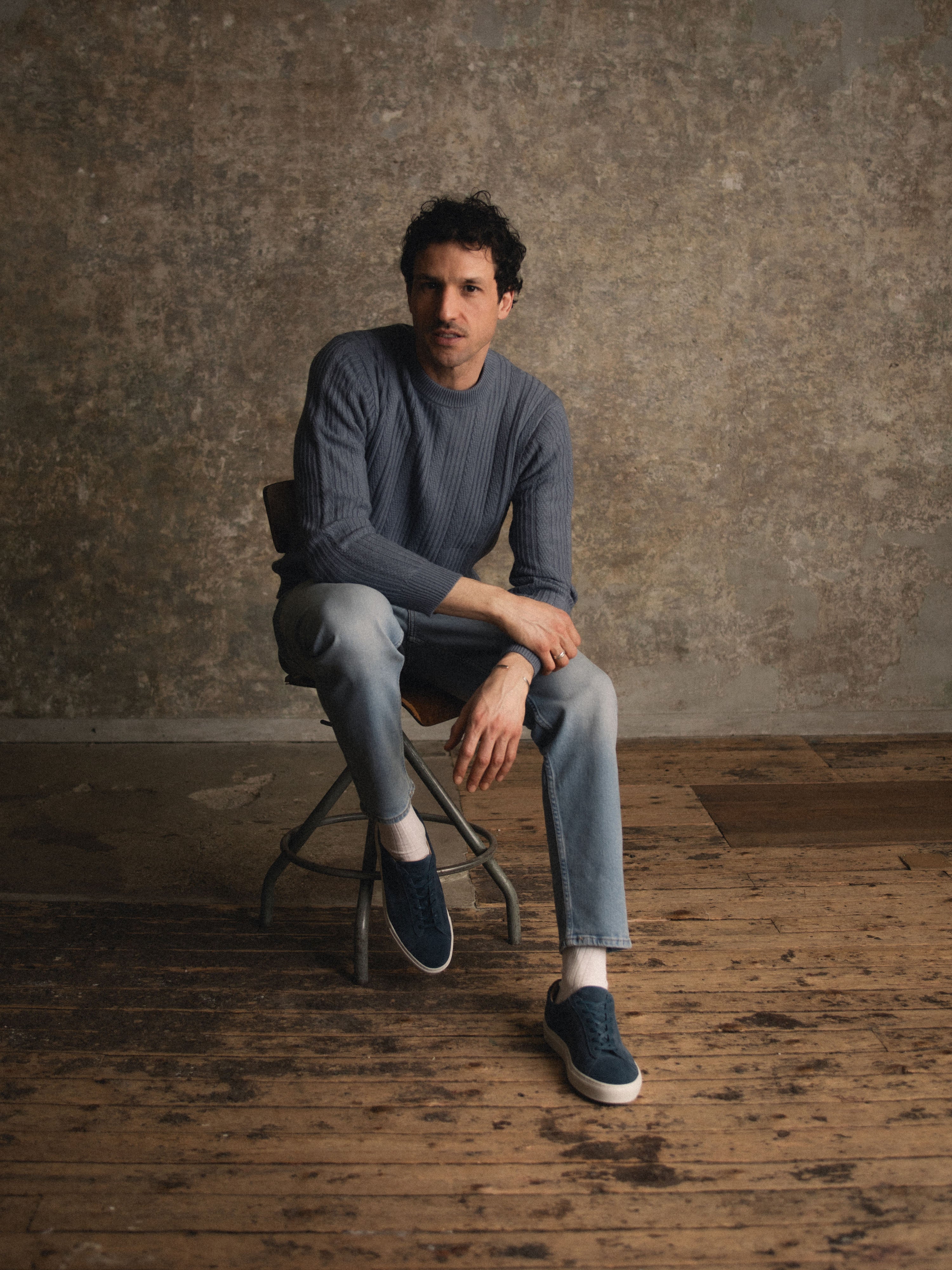 Man sitting on chair wearing jeans, a blue sweater, and dark blue suede sneakers with matching laces and white rubber soles